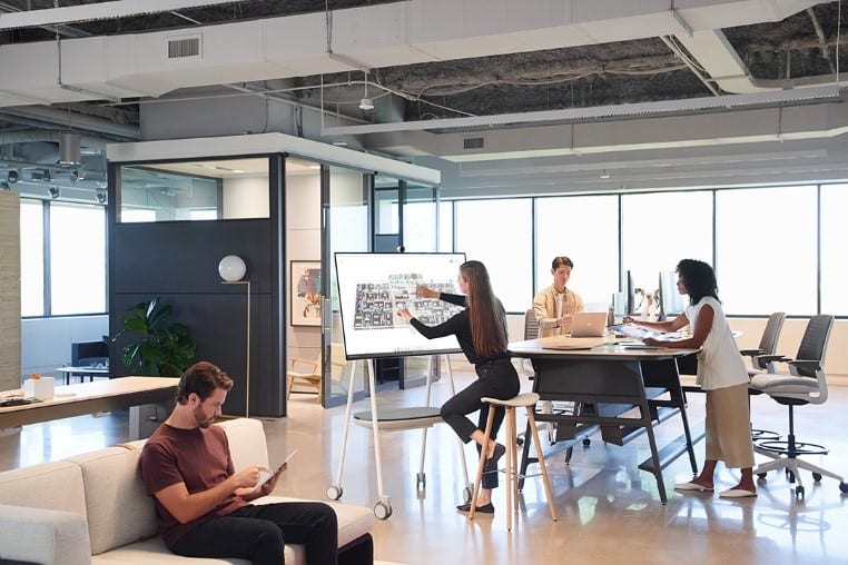 Modern office with large windows: woman writing on whiteboard, two at high table with laptops, man on couch using tablet.