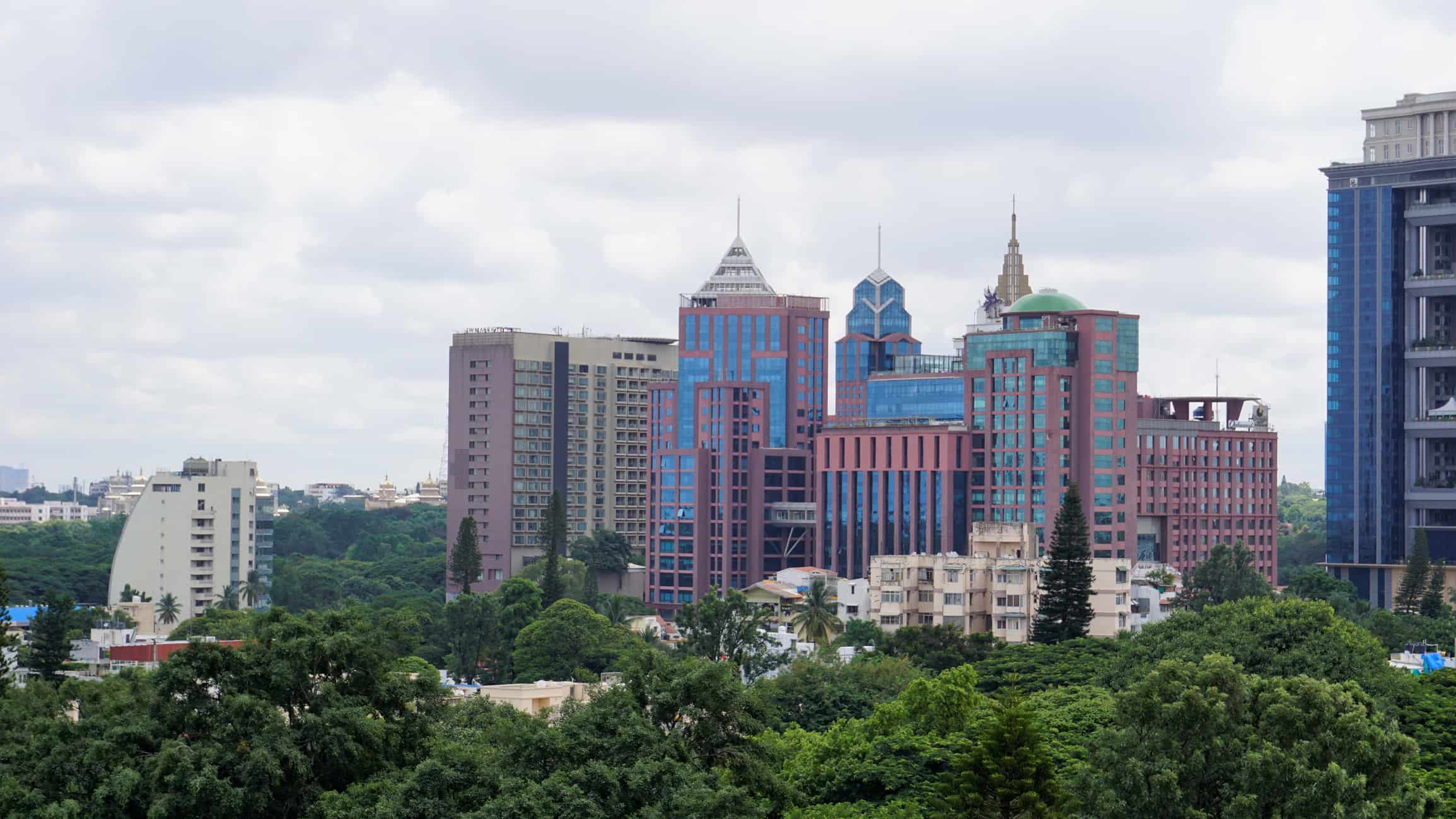 Modern buildings with colorful facades, cloudy sky, green trees in foreground, urban structures in background.