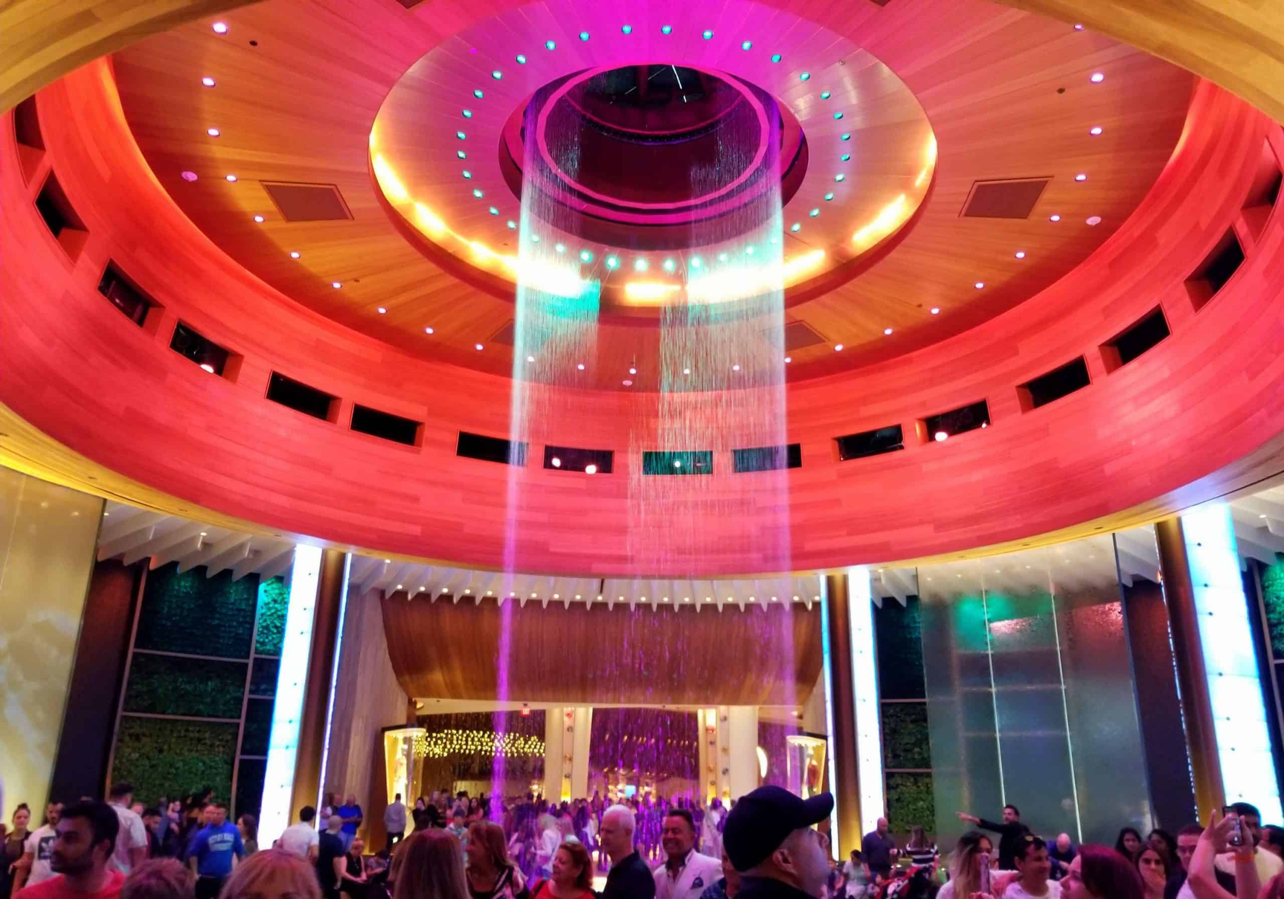 A vibrant indoor scene with a colorful circular ceiling, cascading water, and crowds engaged in conversation and taking photos.
