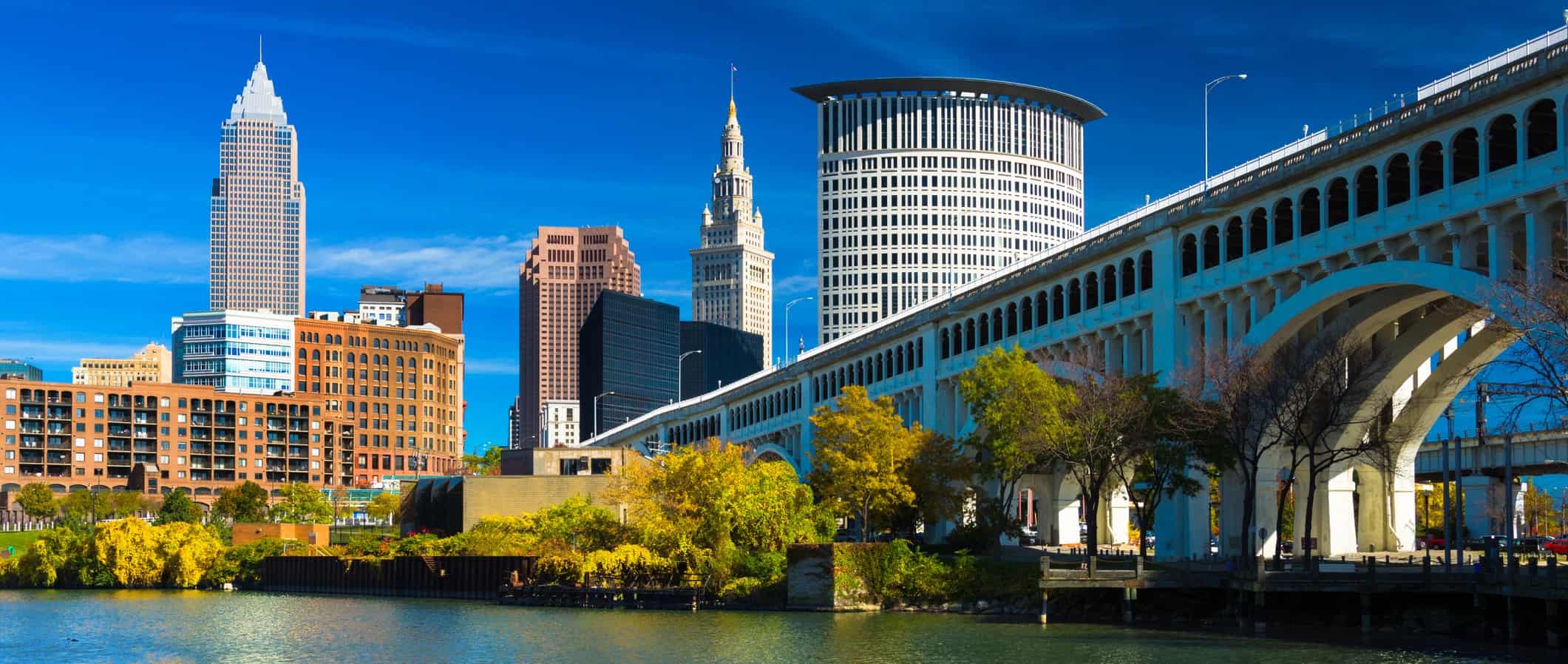 City skyline with a prominent bridge to the right; mixed architecture, river and autumn trees in the foreground, clear blue sky.