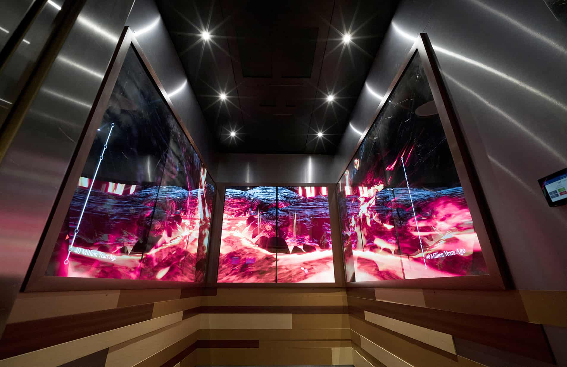 Elevator interior with digital screens displaying abstract pink and blue art; recessed lit ceiling, wood, and metal paneled walls.