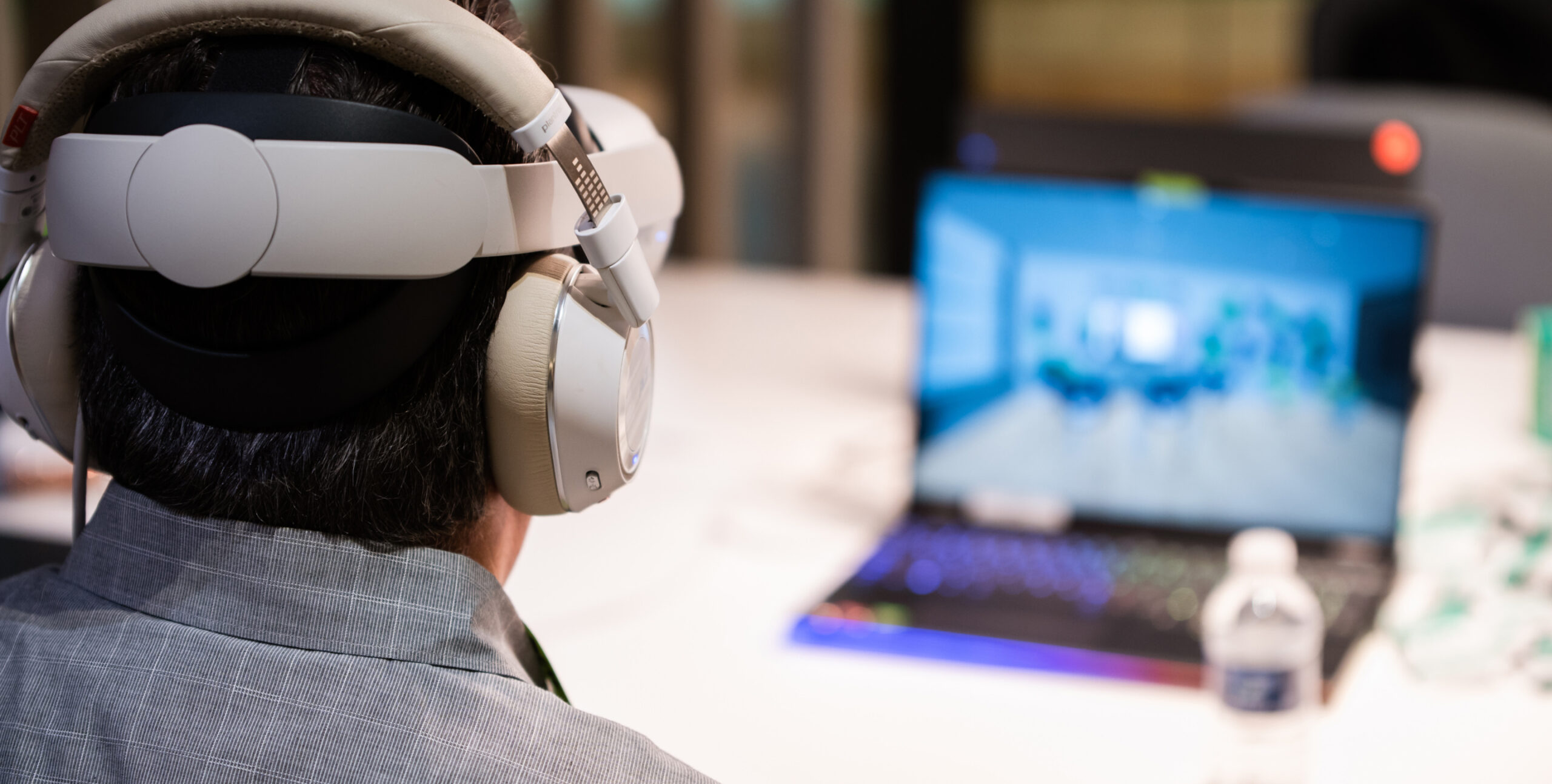 Person with large headphones at desk, open laptop with blurred image, water bottle, device; modern workspace setting.