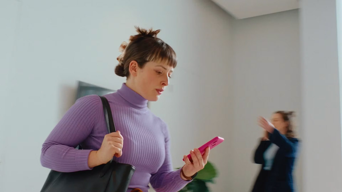 Woman with bun, purple turtleneck, holding pink phone and black bag in bright room; background person blurred, filming with phone.