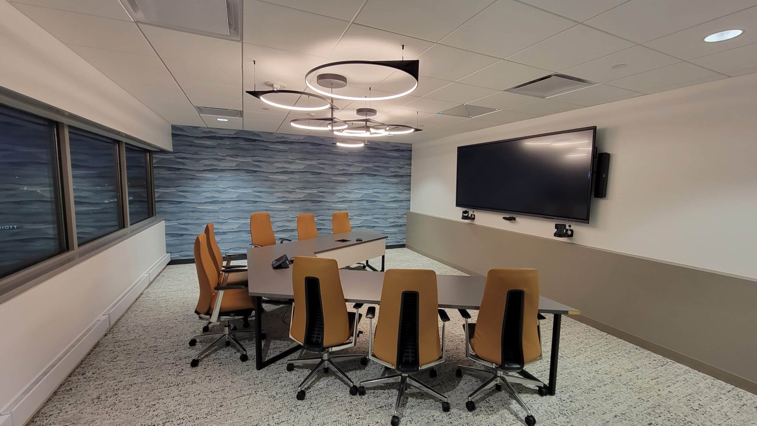 Modern conference room: long table, 8 tan chairs, TV on wave-pattern wall, circular lights, window on left, light carpet.