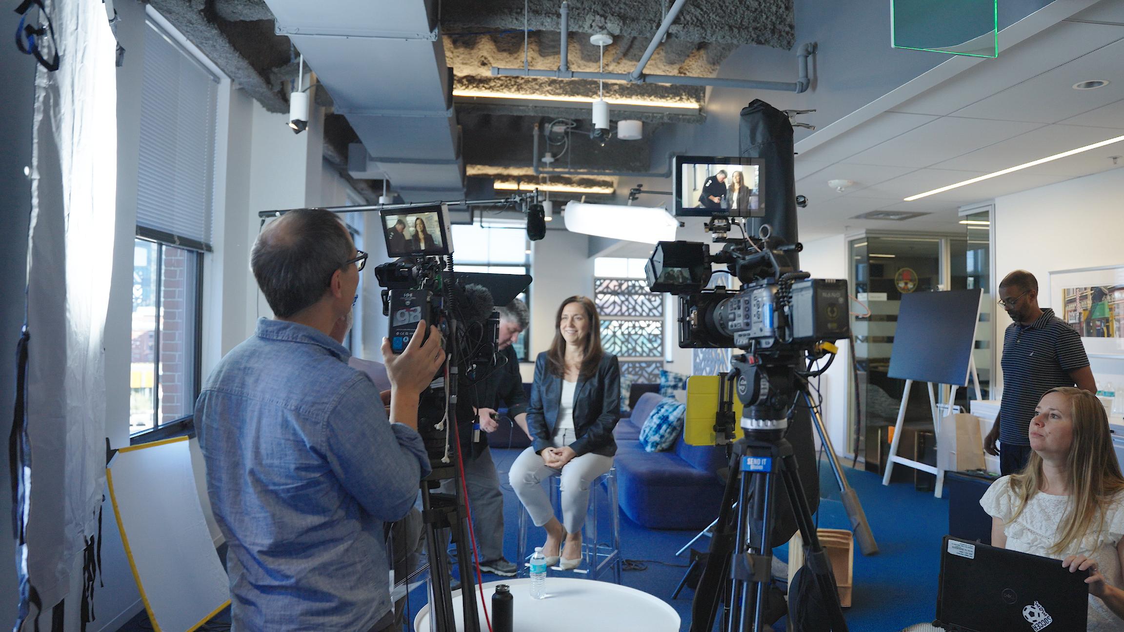 Smiling woman sits in studio surrounded by cameras and crew; natural light streams in, revealing equipment in modern office.