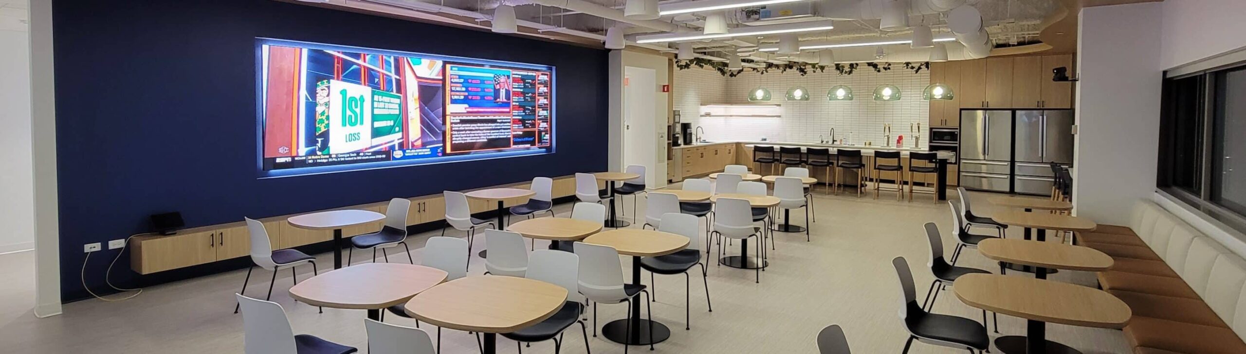 Modern cafeteria: round tables, chairs, large screen on walls, kitchenette with counters, fridges, coffee machine; exposed ducts.