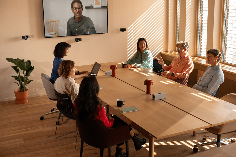 Five people sit around a table, engaged in a video call; room with wooden floors, plant, and filtered sunlight through blinds.