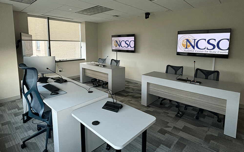 Modern conference room with 3 workstations, ergonomic chairs, computers; NCSC logo on screens; natural light from large left window.