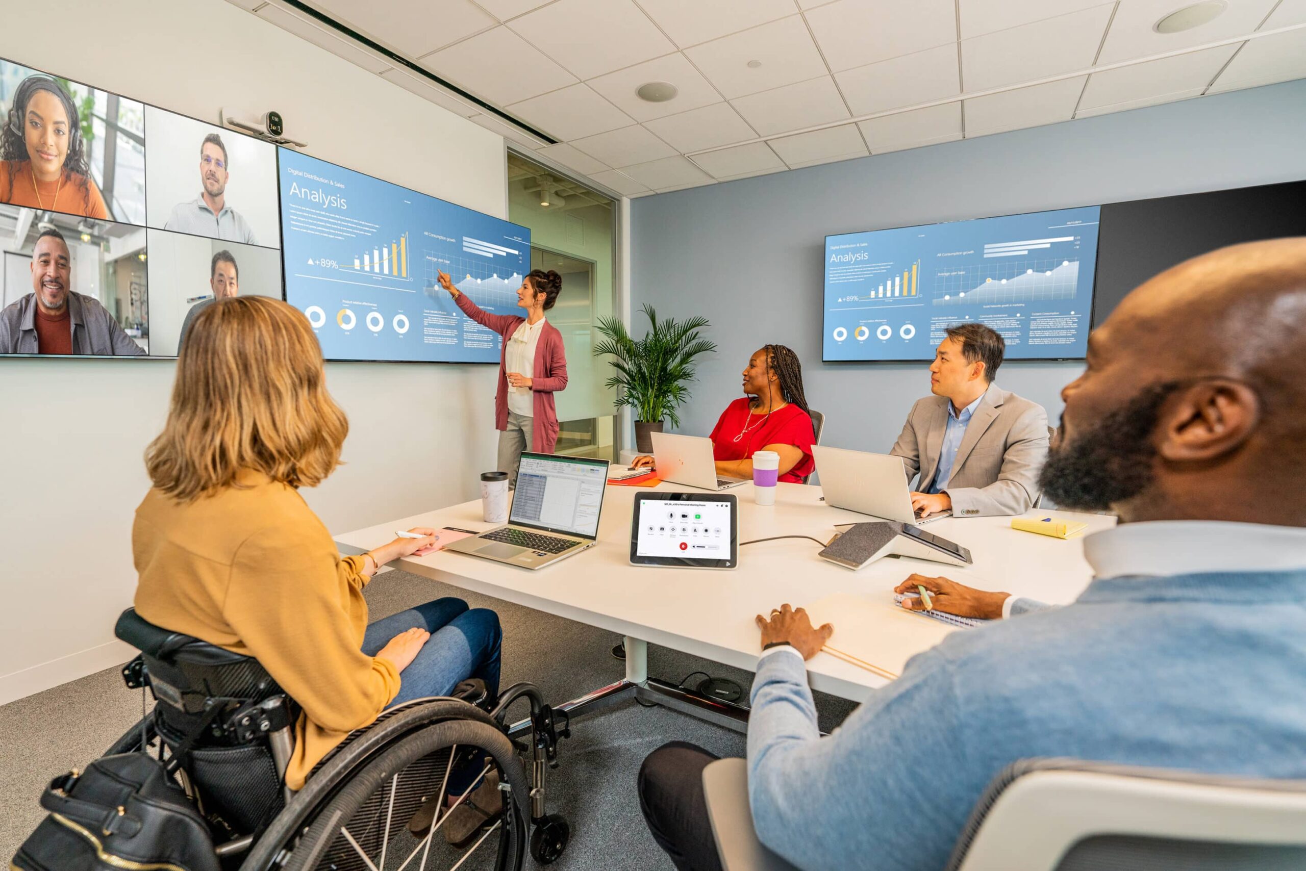 A diverse group in a conference room engaged in a video meeting, with charts on screen and laptops open; a plant in the background.